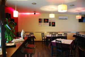 Sala com mesas quadradas, toalhas brancas e trilhos roxos, cadeiras escuras, iluminação pendente e zona de bar com plantas decorativas.