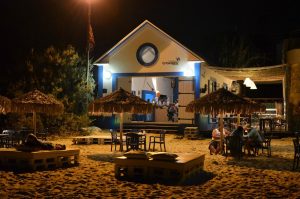 Bar de praia com fachada branca e detalhes azuis, iluminação quente, mesas e cadeiras sobre a areia, pessoas sentadas e guarda‑sóis de colmo num cenário noturno.