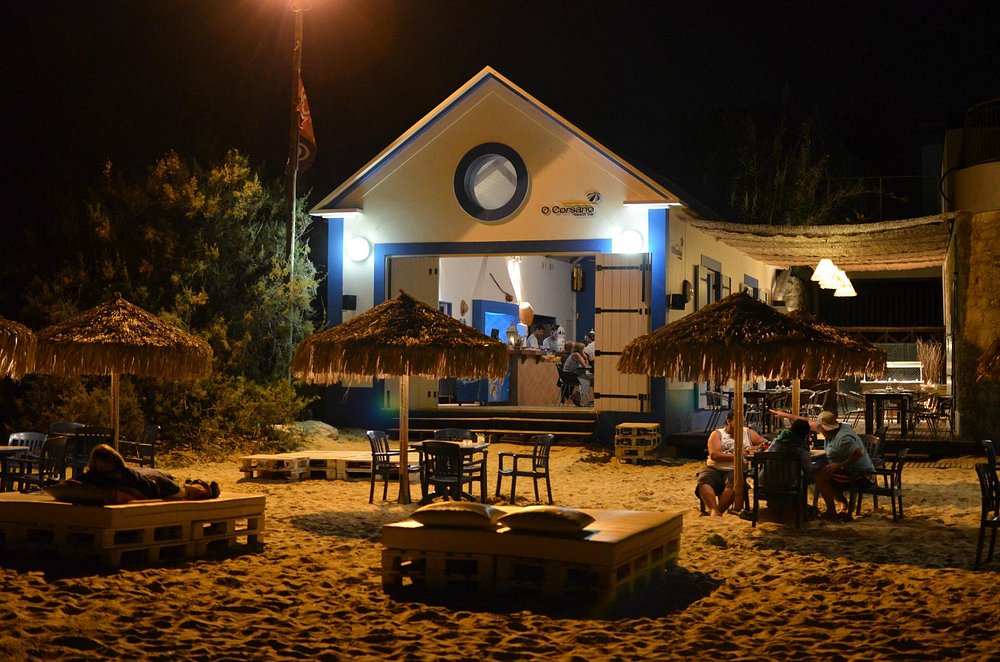 Bar de praia com fachada branca e detalhes azuis, iluminação quente, mesas e cadeiras sobre a areia, pessoas sentadas e guarda‑sóis de colmo num cenário noturno.