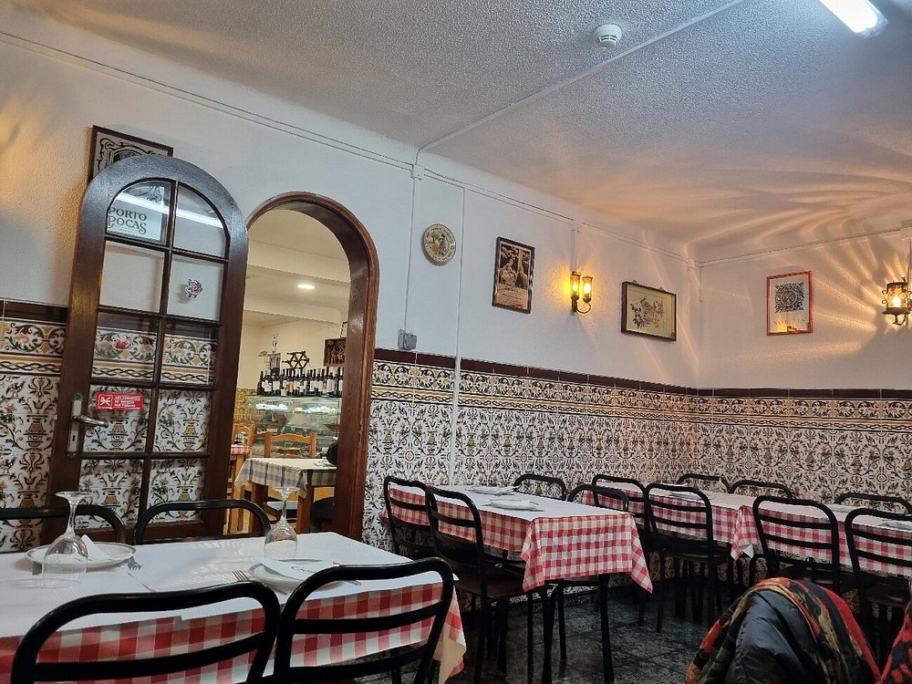 Sala com mesas de toalha axadrezada, cadeiras metálicas, paredes com azulejos decorativos e porta de madeira.