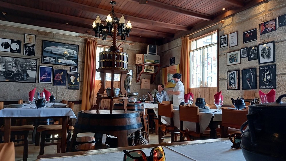 Sala com paredes de pedra, mesas com toalhas brancas, copos de vinho, guardanapos vermelhos e fotografias na parede.