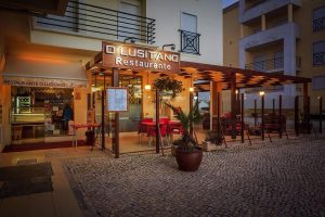 Exterior do Restaurante O Lusitano com pergola de madeira, mesas com toalhas vermelhas e iluminação quente.