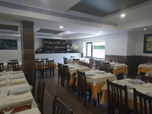 Sala com mesas de madeira cobertas por toalhas creme, copos, pratos e talheres; chão em madeira escura, garrafeira ao fundo e arranjo floral junto à janela.