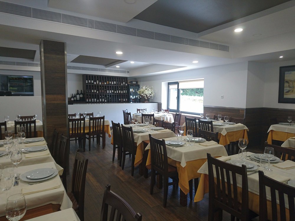 Sala com mesas de madeira cobertas por toalhas creme, copos, pratos e talheres; chão em madeira escura, garrafeira ao fundo e arranjo floral junto à janela.