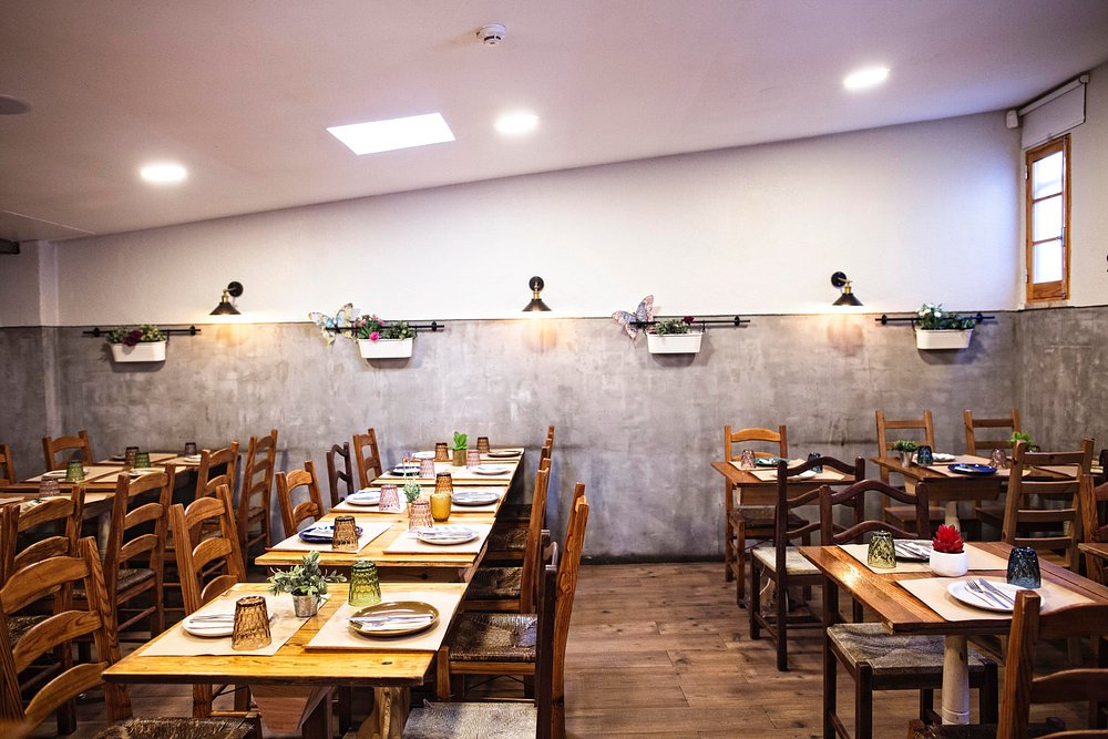 Sala com mesas de madeira preparadas com pratos, talheres, copos e bases individuais, paredes cinzentas com iluminação quente e pequenos vasos decorativos.