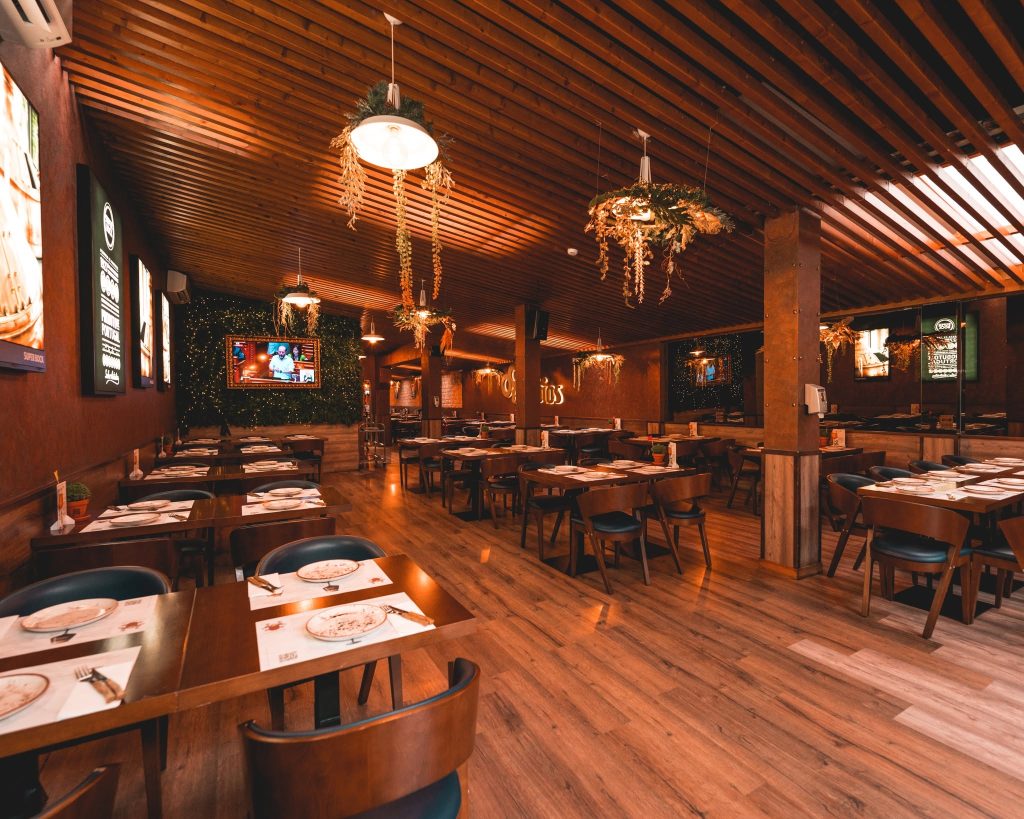 Sala de restaurante com mesas de madeira, teto com ripas, iluminação pendente e decoração com plantas secas.