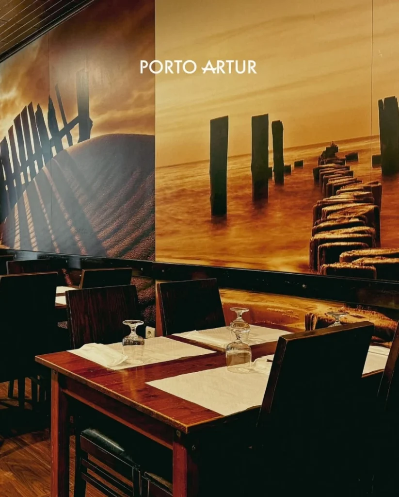 Sala com mesas de madeira, copos virados, cadeiras e mural de praia com tons dourados na parede.