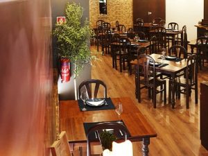 Sala com mesas de madeira, individuais, pratos, copos e talheres; chão em madeira polida, paredes com texturas distintas, planta decorativa e iluminação suave.