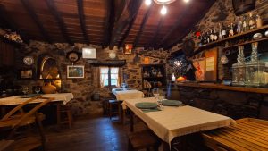 Sala com mesas cobertas por toalhas bege, paredes de pedra, teto com vigas de madeira e bar com garrafas e objetos decorativos.
