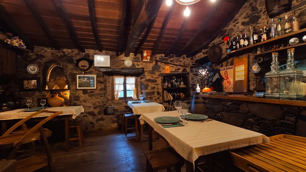 Sala com mesas cobertas por toalhas bege, paredes de pedra, teto com vigas de madeira e bar com garrafas e objetos decorativos.