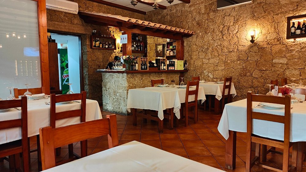 Sala com mesas quadradas cobertas por toalhas brancas, cadeiras de madeira e paredes em pedra.