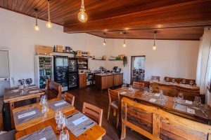 Sala com mesas e cadeiras de madeira, copos e talheres dispostos, teto em madeira com lâmpadas pendentes, balcão ao fundo com garrafeira, frigoríficos e prateleiras com produtos.
