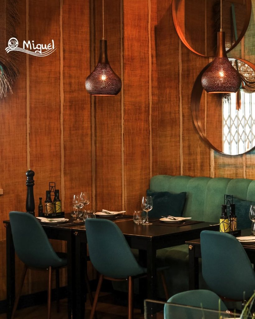 Sala com mesas de madeira escuras, cadeiras azul‑petróleo, banco almofadado, painéis de madeira na parede, candeeiros metálicos suspensos e espelhos decorativos.