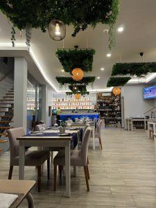 Sala com mesas alinhadas com toalhas azul‑escuro, pratos, talheres e copos invertidos, cadeiras bege, teto com painéis de plantas suspensas e candeeiros, bar iluminado a azul ao fundo e escadas laterais.