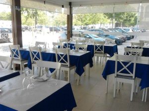 Sala com mesas cobertas por toalhas azuis e brancas, copos virados, guardanapos dobrados, cadeiras brancas e janelas amplas com vista exterior.