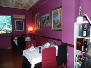 Sala com paredes roxas decoradas com quadros, mesas com toalhas brancas, copos de vinho, cadeiras com capas e prateleira com garrafas e objetos decorativos.