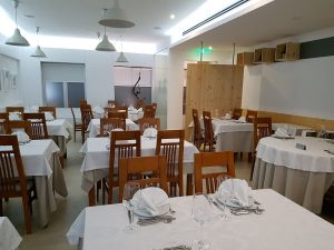 Sala de restaurante com mesas de madeira cobertas por toalhas brancas, cadeiras de madeira, candeeiros suspensos e paredes claras com poucos quadros.