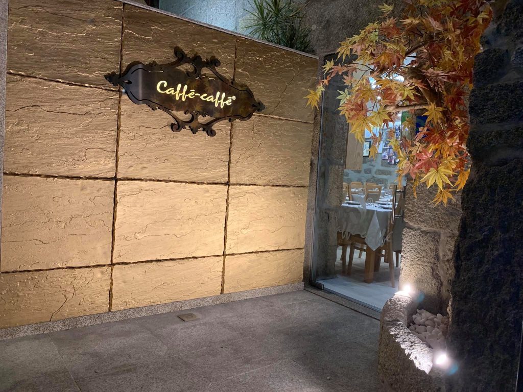 Entrada com letreiro “Caffé‑caffé” em moldura decorativa, parede de pedra iluminada, folhas decorativas e acesso à sala com mesas preparadas.