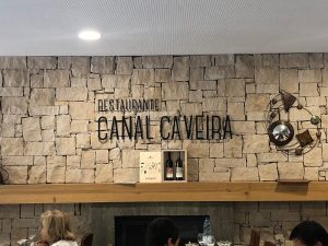 Parede de pedra com letreiro “Restaurante Canal Caveira”, caixa com garrafas e peça metálica decorativa.