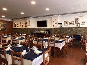 Sala com mesas de madeira cobertas por toalhas brancas e caminhos azuis, paredes com quadros e utensílios antigos, e garrafeira lateral.