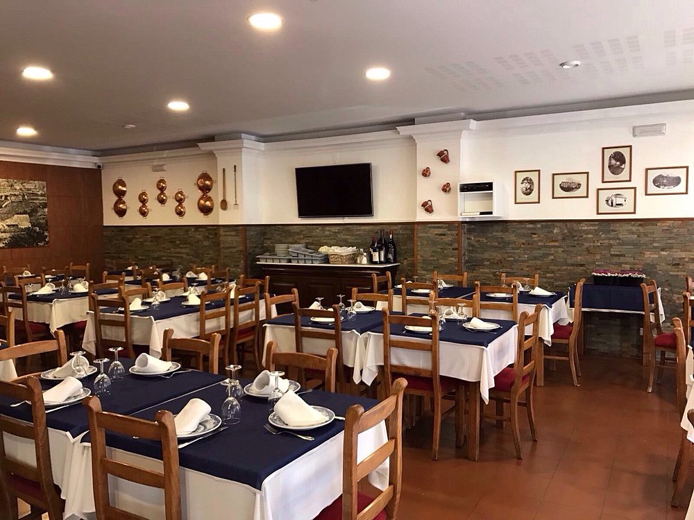 Sala com mesas de madeira cobertas por toalhas brancas e caminhos azuis, paredes com quadros e utensílios antigos, e garrafeira lateral.