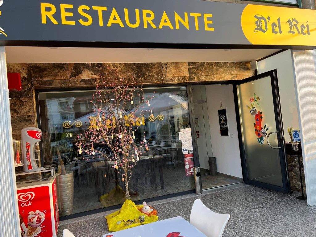 Entrada do restaurante com fachada moderna, mesas exteriores, cadeiras brancas, decoração festiva e equipamento de gelados junto à porta envidraçada.