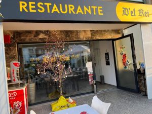 Entrada do restaurante com fachada moderna, mesas exteriores, cadeiras brancas, decoração festiva e equipamento de gelados junto à porta envidraçada.