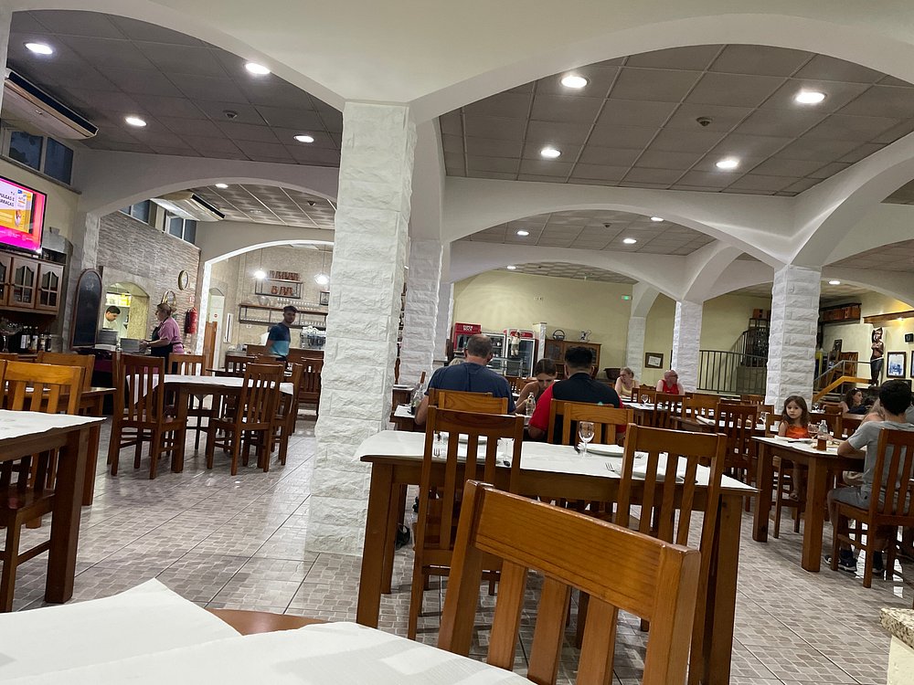 Sala com mesas de madeira, colunas brancas, pessoas a comer e balcão ao fundo.