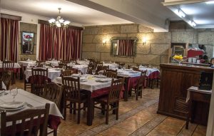 Sala com mesas de madeira cobertas com toalhas brancas e vermelhas, paredes de pedra, espelho grande e balcão com fruta e máquina de café.