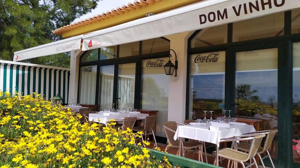Esplanada com mesas de toalha branca, copos e cadeiras, cobertura com o nome “Dom Vinho”, janelas com sinalética e canteiro de flores amarelas em primeiro plano.