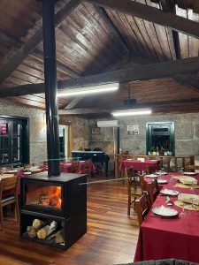 Sala de restaurante com chão e teto de madeira, paredes de pedra, fogão a lenha aceso ao centro, mesas com toalhas vermelhas e decoração tradicional.
