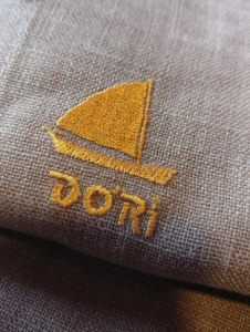 Tecido bege com bordado amarelo representando barco de vela e palavra “DORI” por baixo.