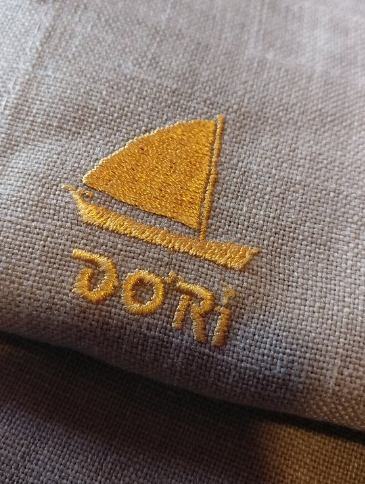 Tecido bege com bordado amarelo representando barco de vela e palavra “DORI” por baixo.