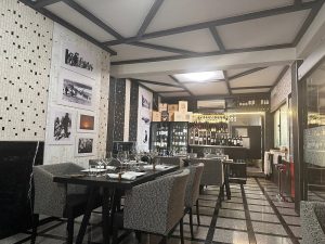 Sala com teto geométrico, mesas com copos e guardanapos, paredes com mosaicos e fotografias, e bar com garrafas e copos suspensos.