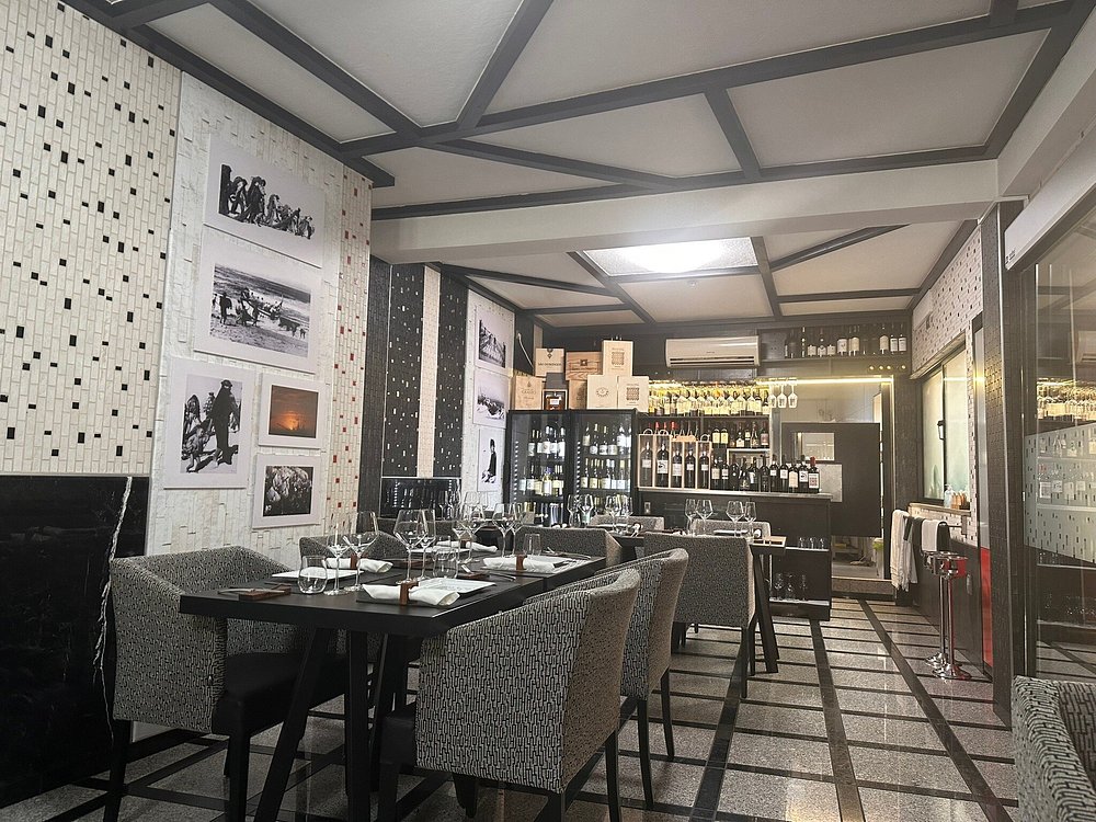 Sala com teto geométrico, mesas com copos e guardanapos, paredes com mosaicos e fotografias, e bar com garrafas e copos suspensos.