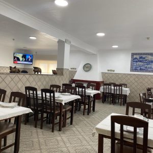 Sala com mesas de madeira cobertas por toalhas brancas, paredes com azulejos, televisão ligada, mural decorativo e pequenas figuras de animais sobre a divisória.
