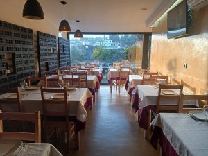 Sala de restaurante com mesas de madeira cobertas por toalhas brancas e saias vermelhas, prateleiras com garrafas, paredes douradas e janelas amplas com vista para árvores.