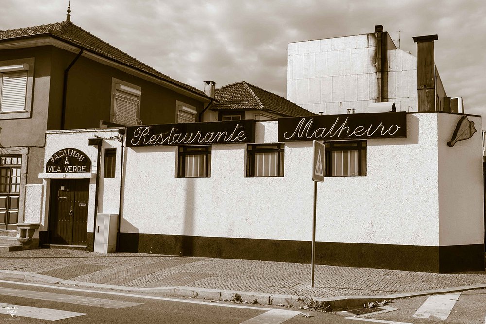 Fachada de restaurante com placa “Restaurante Malheiro”, paredes claras, telhado de telha, porta lateral com letreiro “Bacalhau à Vila Verde” e passadeira de peões à frente.