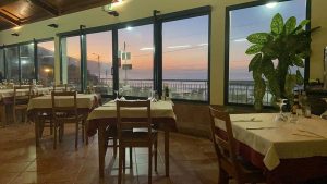 Sala de restaurante com mesas montadas e grandes janelas que mostram o pôr do sol sobre o mar.