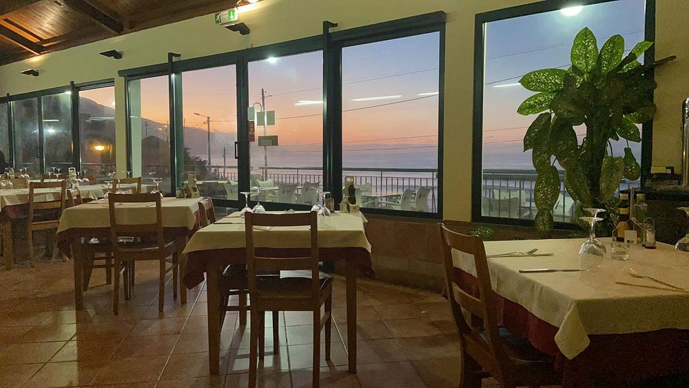 Sala de restaurante com mesas montadas e grandes janelas que mostram o pôr do sol sobre o mar.