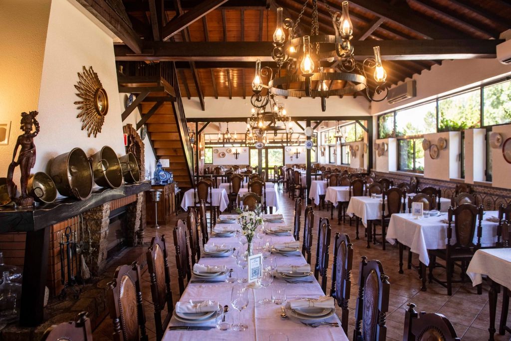 Sala com mesas de madeira cobertas por toalhas brancas, lareira decorada, teto alto com vigas e iluminação quente.