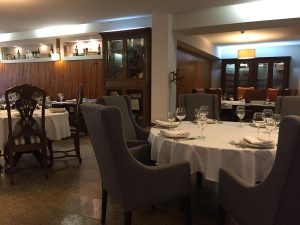 Sala de restaurante com mesas redondas cobertas por toalhas brancas, copos de vinho, cadeiras estofadas e prateleiras de madeira com garrafas e objetos decorativos.