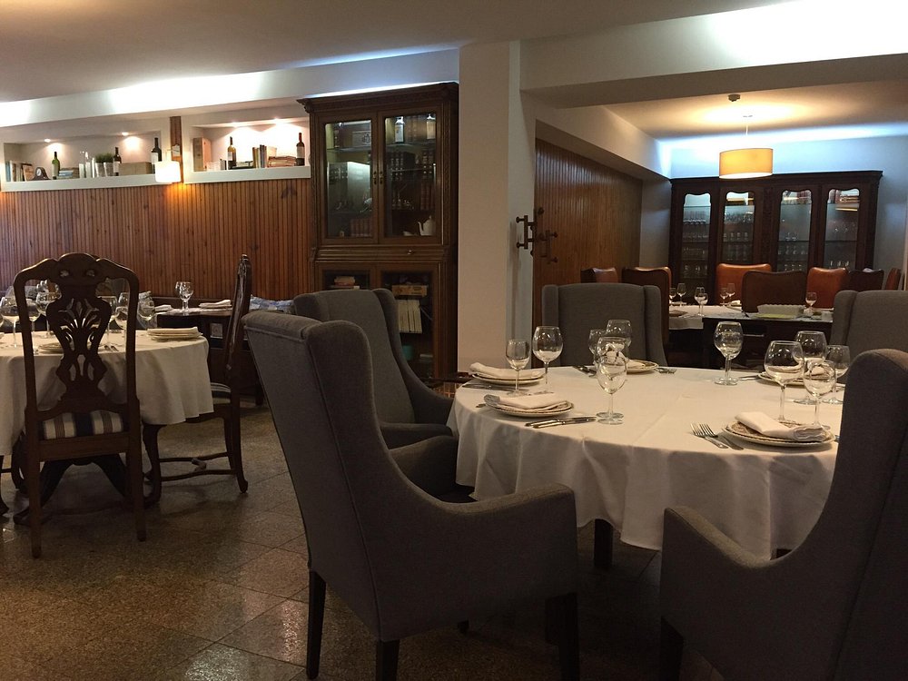 Sala de restaurante com mesas redondas cobertas por toalhas brancas, copos de vinho, cadeiras estofadas e prateleiras de madeira com garrafas e objetos decorativos.
