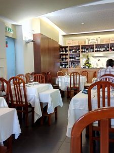 Sala de restaurante com mesas cobertas por toalhas brancas, copos de vinho, cadeiras de madeira e prateleiras com garrafas ao fundo, com funcionário atrás do balcão.