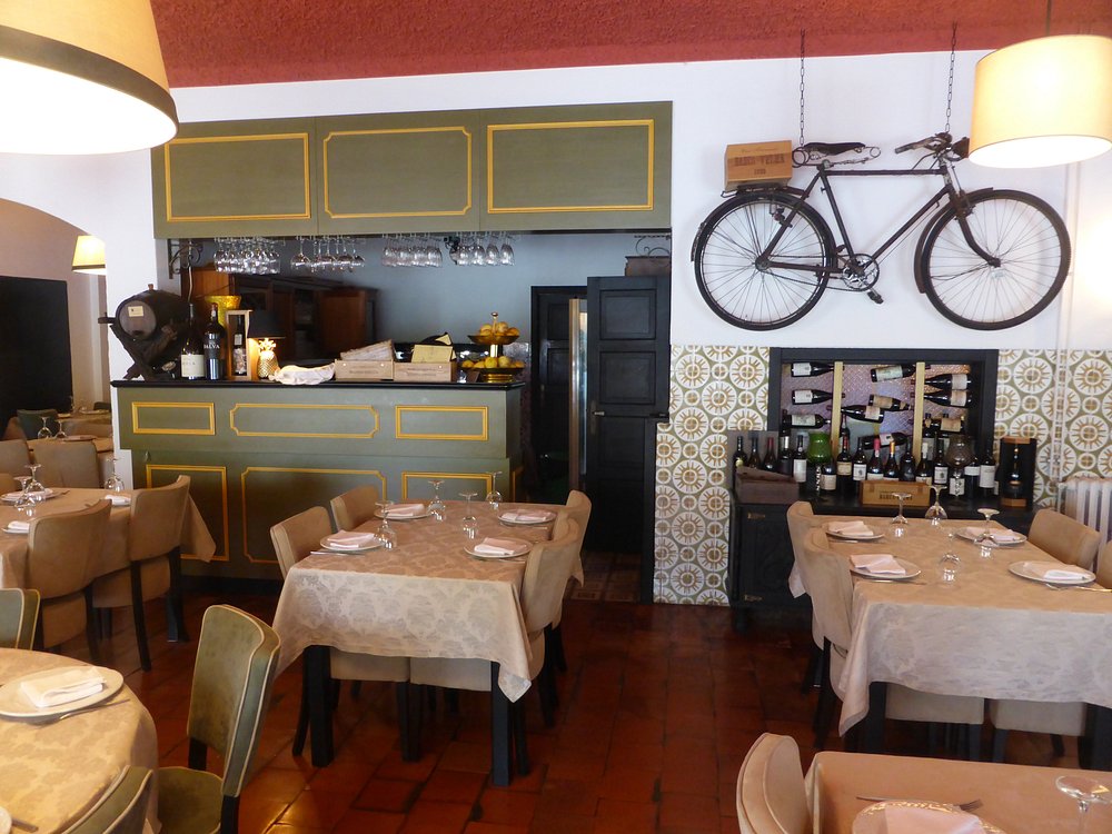 Sala com mesas de toalha bege, chão em tijoleira, paredes com azulejos decorativos, bicicleta pendurada, garrafeira, balcão verde com copos suspensos e iluminação quente.