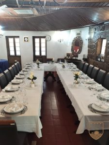 Sala de refeição com mesas em formato U, toalhas brancas, pratos, talheres, copos, guardanapos enrolados, centros de mesa com rosas brancas, cadeiras escuras, chão em tijoleira, janelas com molduras de madeira, espelho decorativo e garrafas de vinho numa mesa lateral.