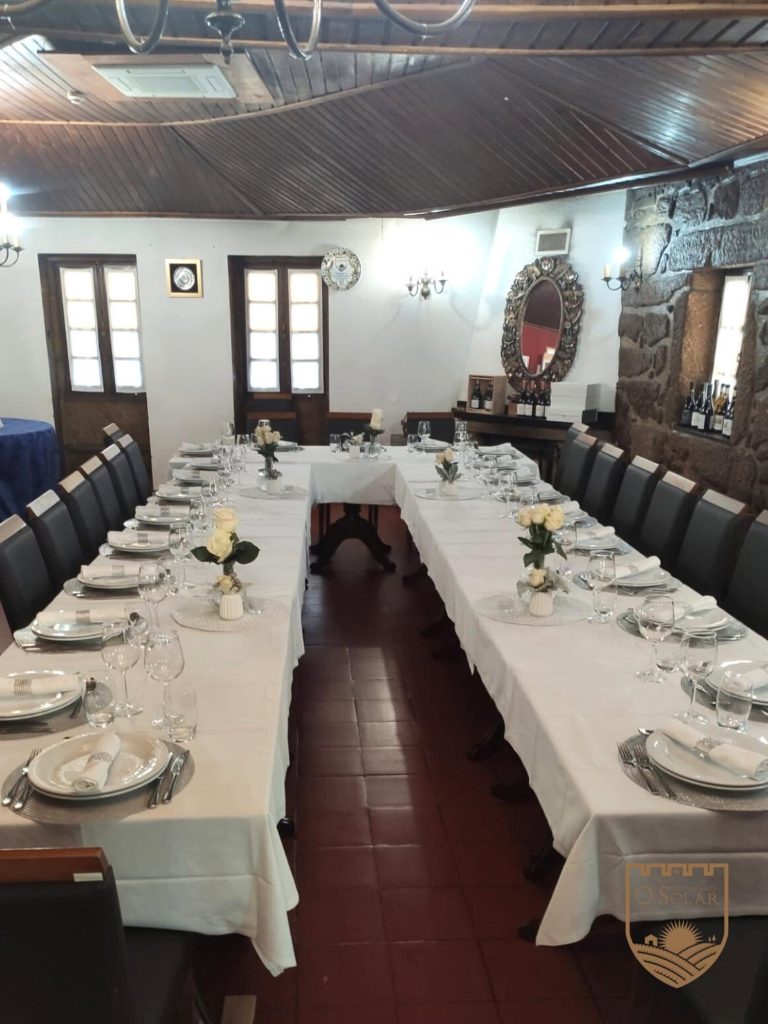 Sala de refeição com mesas em formato U, toalhas brancas, pratos, talheres, copos, guardanapos enrolados, centros de mesa com rosas brancas, cadeiras escuras, chão em tijoleira, janelas com molduras de madeira, espelho decorativo e garrafas de vinho numa mesa lateral.