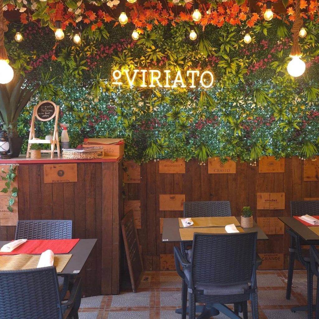 Sala com parede coberta por plantas e flores, letreiro luminoso “VIRIATO”, lâmpadas pendentes, mesas com bases individuais e cadeiras entrançadas, além de zona lateral com cestos e garrafas.