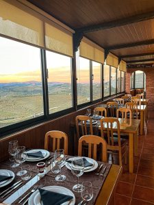 Sala de restaurante com mesas de madeira preparadas com pratos, copos e guardanapos escuros, junto a janelas amplas que mostram paisagem de vinhas ao entardecer.
