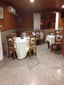 Sala de restaurante com mesas redondas cobertas por toalhas brancas, cadeiras de madeira e janela com cortinas claras.